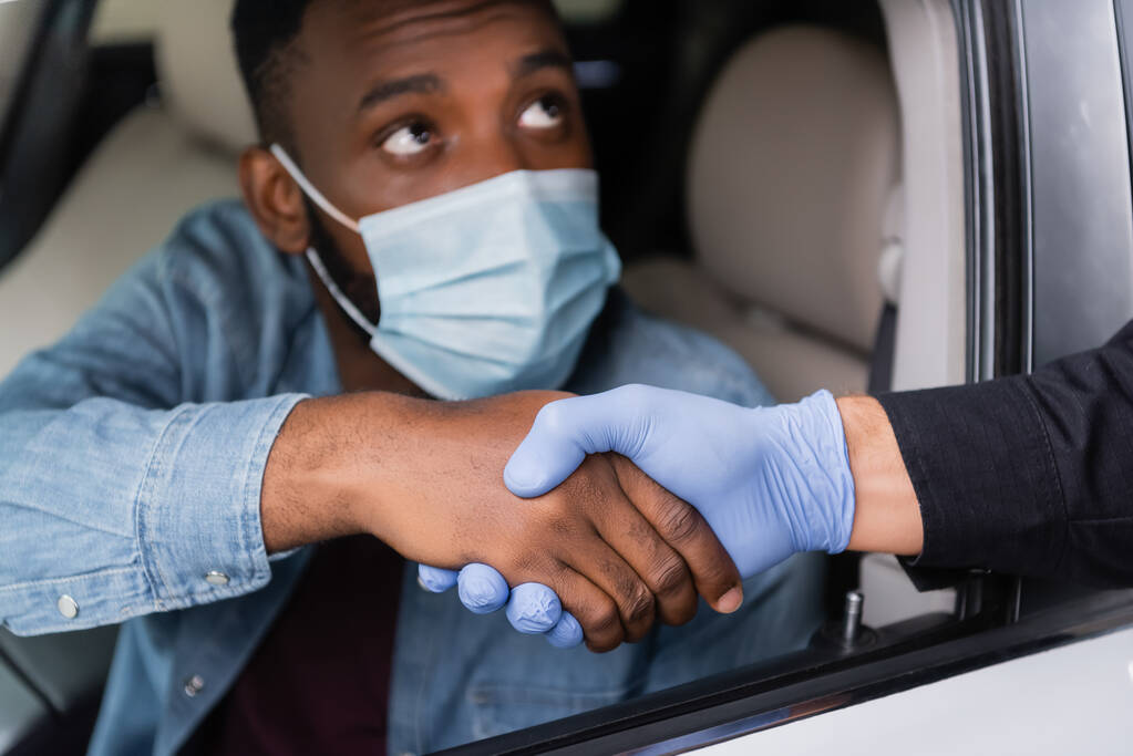 Police officer in latex glove handshaking with african american driver in medical mask on blurred background in car  - Photo, Image