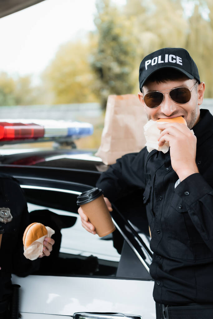 Oficial de policía comiendo hamburguesa y sosteniendo café para ir cerca de colega y coche en un fondo borroso  - Foto, Imagen