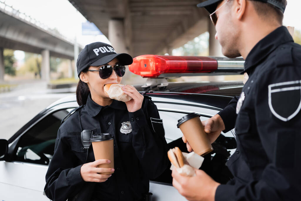 Mujer policía comiendo hamburguesa y sosteniendo café para ir cerca de colega y coche de policía en un fondo borroso  - Foto, Imagen