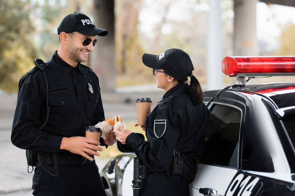 Un policier souriant tenant un café à emporter et un hamburger près d'un collègue pendant le déjeuner près de la voiture à l'extérieur  - Photo, image