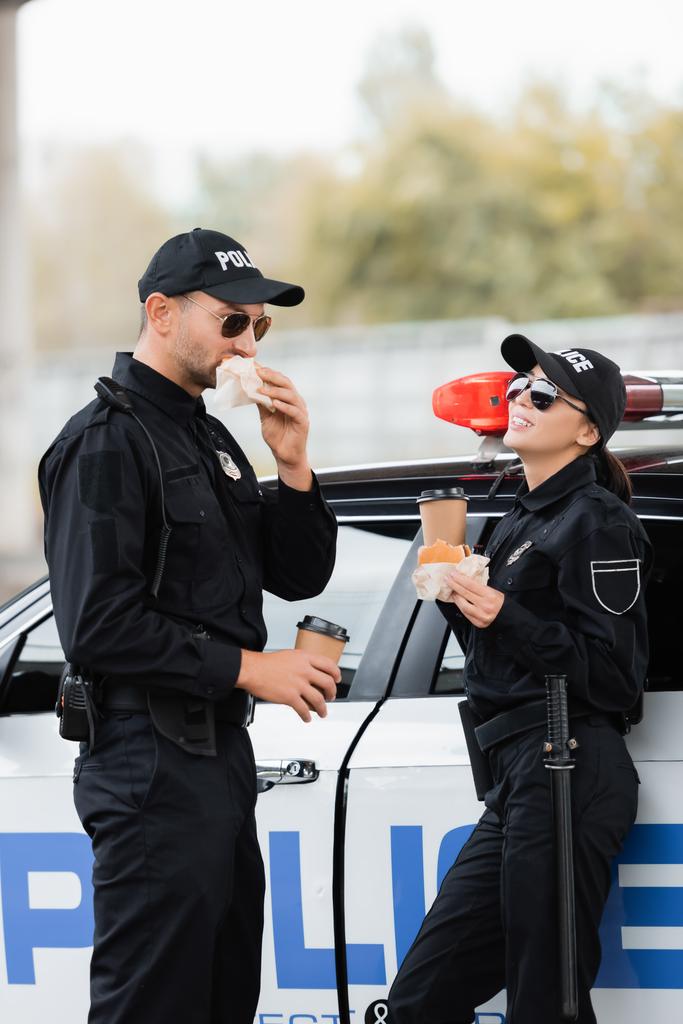Mujer policía sonriente en gafas de sol sosteniendo hamburguesa y café para ir cerca de colega comer y coche al aire libre  - Foto, imagen