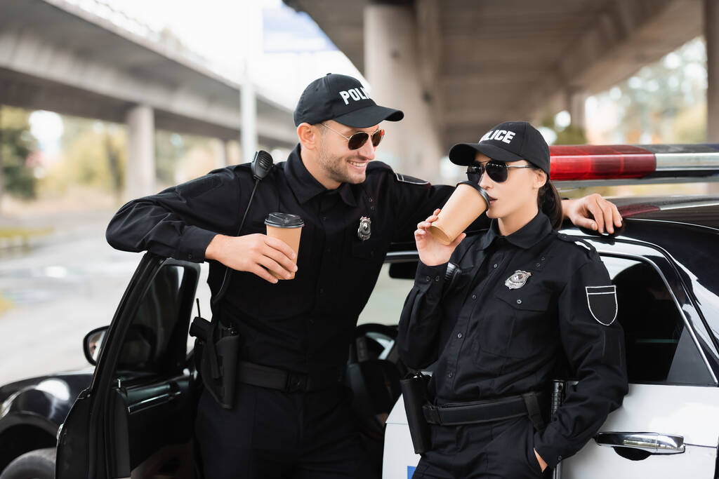 Усміхнений офіцер поліції в сонцезахисних окулярах дивиться на колегу, яка п'є каву, щоб підійти до машини на відкритому повітрі
  - Фото, зображення