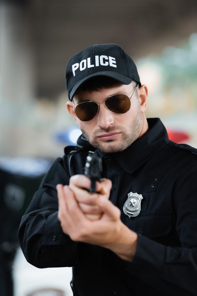 Police officer in uniform and sunglasses holding gun on blurred foreground outdoors  - Photo, Image