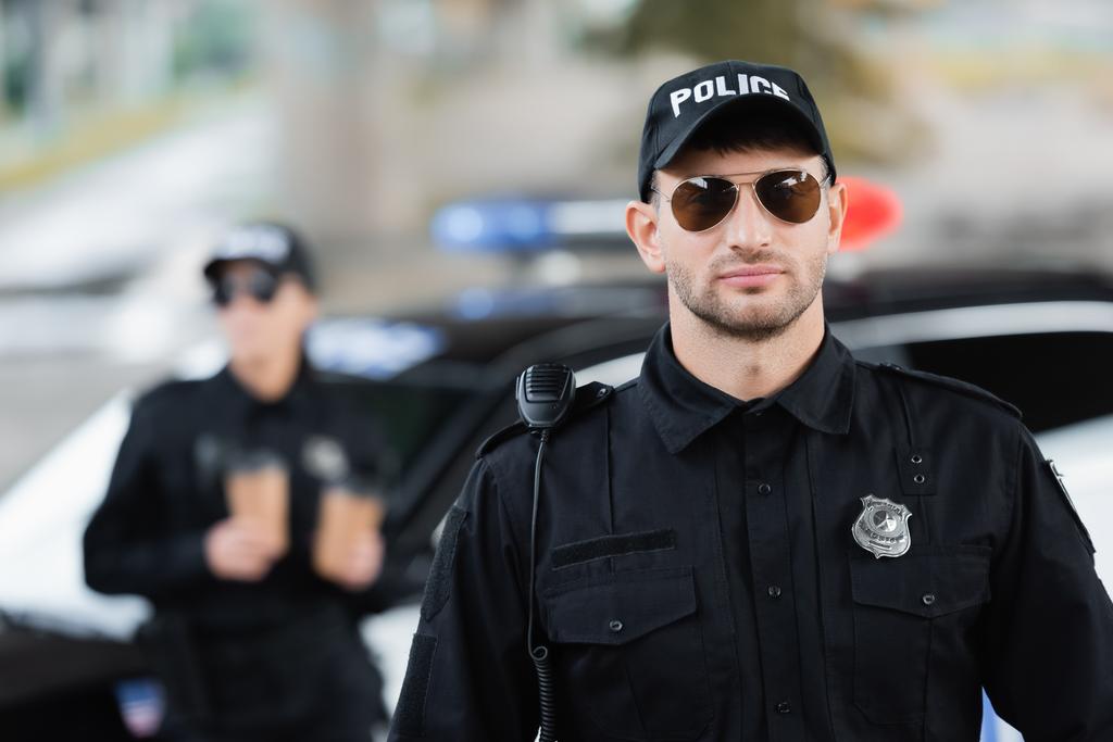 Politieagent met een zonnebril in de buurt van collega en auto op wazige achtergrond buiten  - Foto, afbeelding