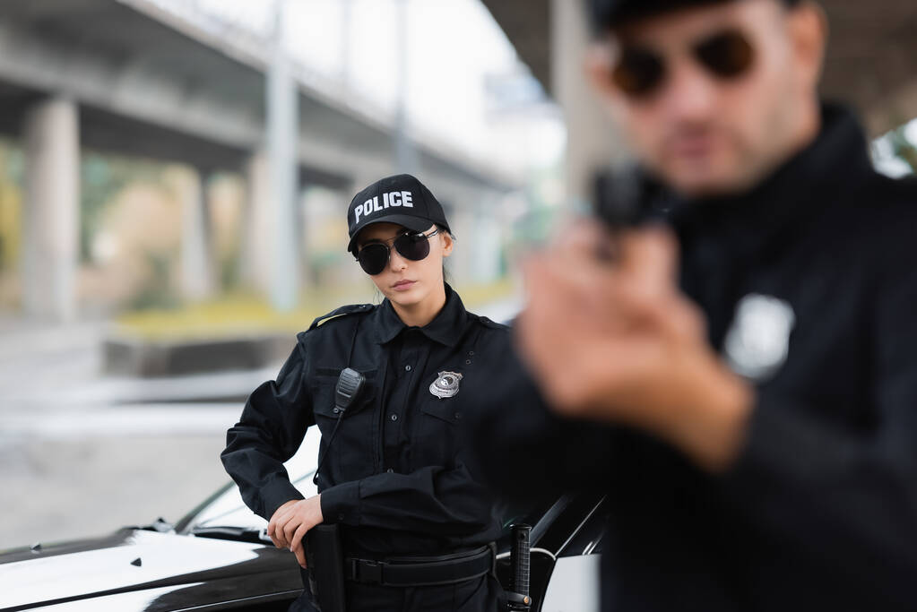 Policjantka w okularach przeciwsłonecznych biorąc pistolet w pobliżu samochodu i kolegi na niewyraźnym pierwszym planie na świeżym powietrzu  - Zdjęcie, obraz