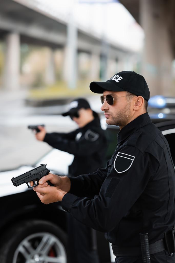 Güneş gözlüklü bir polis memuru iş arkadaşının yanında silah tutuyor ve arka planı bulanık.  - Fotoğraf, Görsel