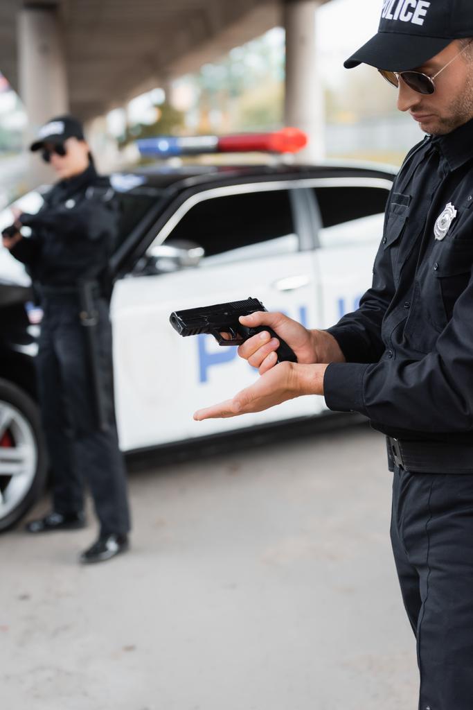 Αξιωματικός της αστυνομίας φορτώνει όπλο κοντά στο συνάδελφο και το αυτοκίνητο σε θολή φόντο σε αστικό δρόμο  - Φωτογραφία, εικόνα