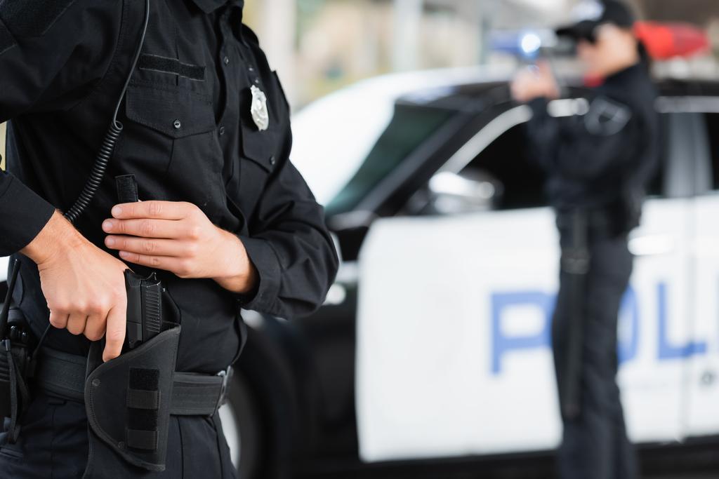 Полицейский взял пистолет из кобуры рядом с коллегой и машиной на размытом фоне  - Фото, изображение