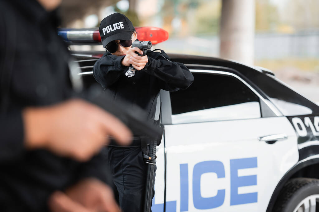 Policjantka w okularach przeciwsłonecznych trzymająca broń w pobliżu kolegi na zamglonym pierwszym planie i w samochodzie na zewnątrz  - Zdjęcie, obraz