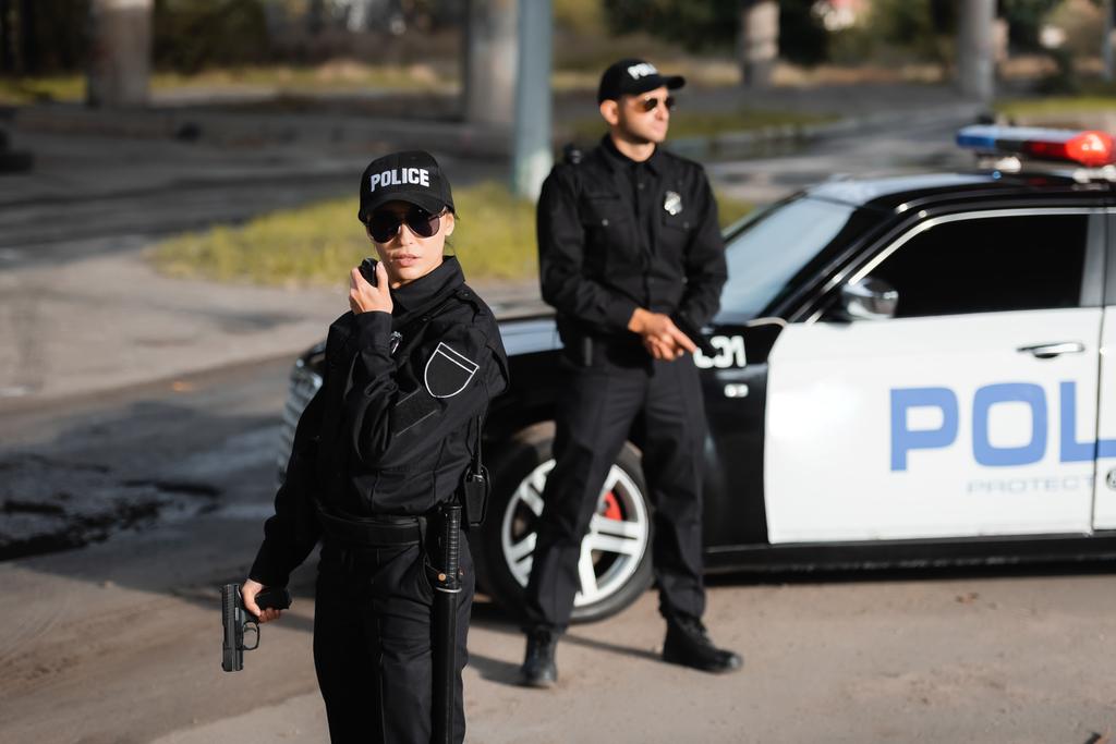 Mujer policía con pistola usando walkie talkie cerca de colega y coche de policía sobre fondo borroso en la calle urbana  - Foto, Imagen