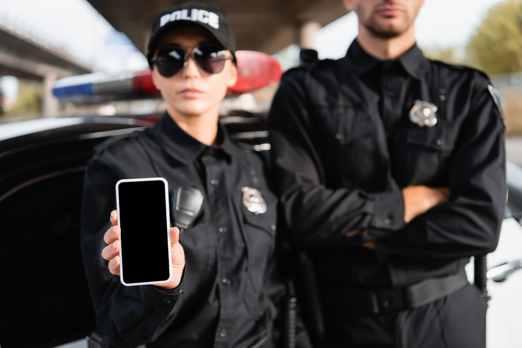 Smartphone con schermo bianco in mano della poliziotta vicino collega e auto su sfondo sfocato  - Foto, immagini