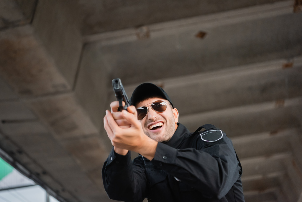Χαμηλή γωνία άποψη του χαρούμενου αστυνομικού σε γυαλιά ηλίου κρατώντας όπλο σε θολή πρόσοψη σε εξωτερικούς χώρους  - Φωτογραφία, εικόνα
