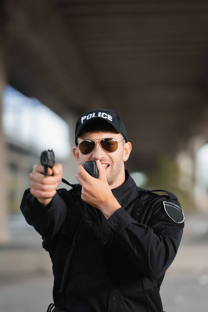Policial em óculos de sol usando walkie talkie e segurando arma em primeiro plano borrado ao ar livre  - Foto, Imagem
