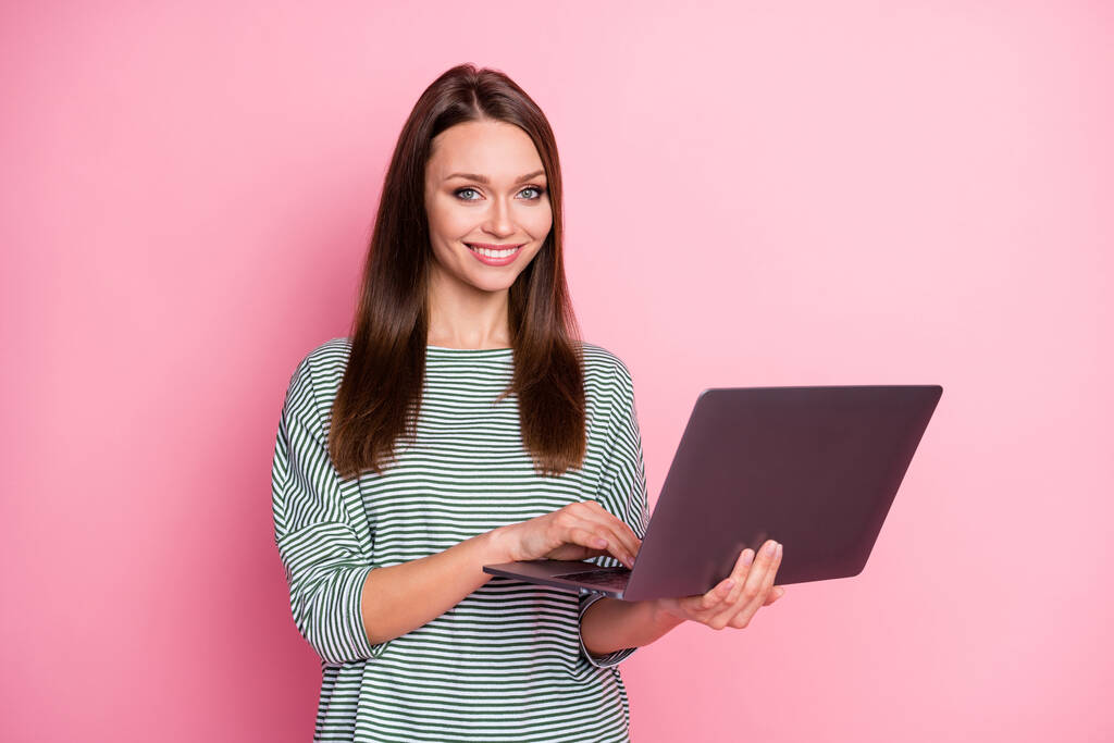 Фото оптимистичной брюнетки длинные волосы женщина работает ноутбук носить повседневные штаны футболки изолированы на пастельно-розовый цвет фона - Фото, изображение