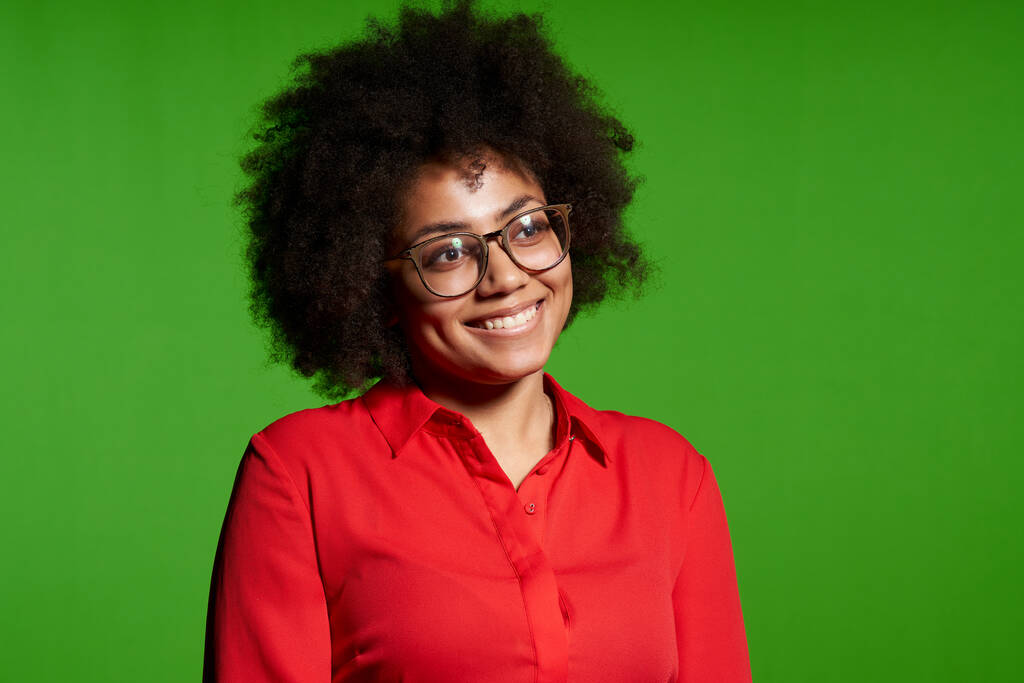Primer plano de la joven afroamericana sonriente con gafas y camisa roja mirando hacia un lado en el espacio de copia en blanco, sobre fondo verde - Foto, imagen