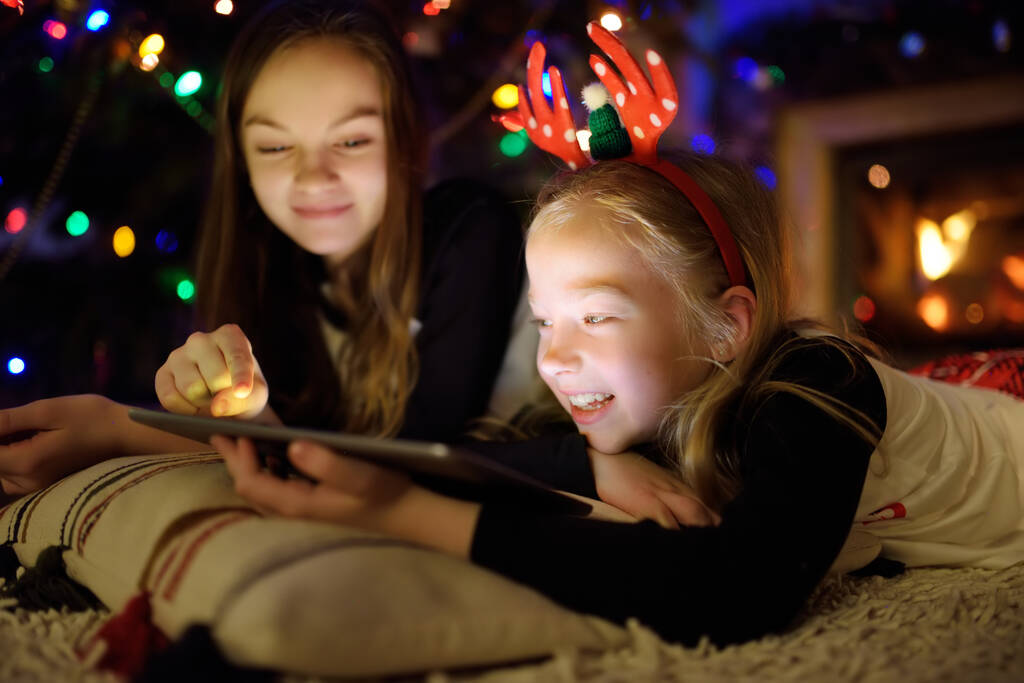 Két aranyos fiatal nővér használ egy tabletta pc otthon egy kandalló meleg és hangulatos nappali karácsony este. Téli este otthon a családdal és a gyerekekkel. - Fotó, kép