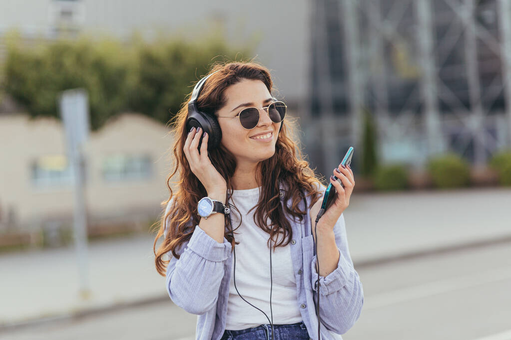 Молодая симпатичная студентка, ждет такси и слушает музыку из телефона на больших наушниках - Фото, изображение