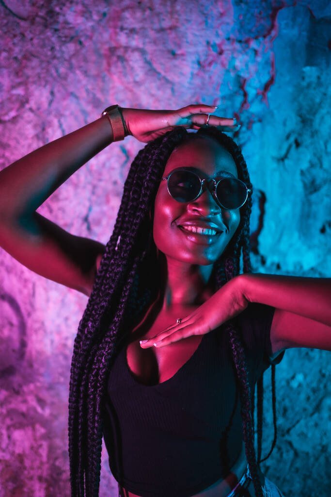 Lifestyle, fille noire avec de longues tresses, des lunettes de soleil et un t-shirt noir. Portrait avec néons roses et bleus, photographie urbaine d'une danseuse piège - Photo, image