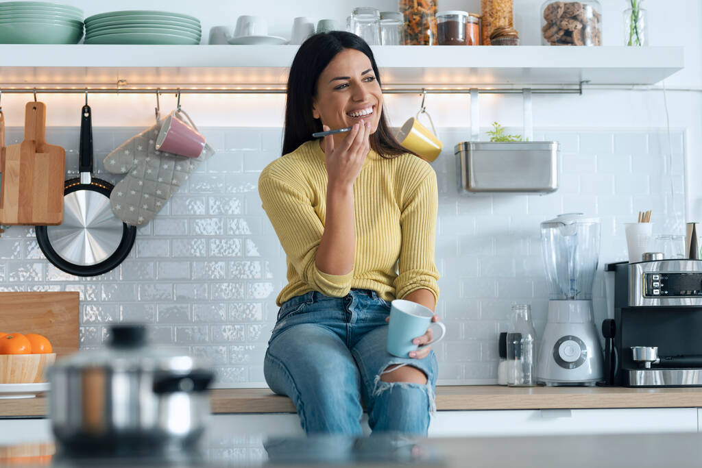 Shot van aantrekkelijke jonge vrouw het maken van een voice message met smart phone terwijl ontspannen drinken koffie zit op de keuken thuis. - Foto, afbeelding