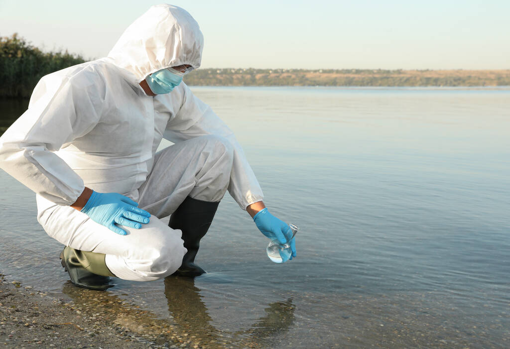 分析のために川からサンプルを取ってフローレンスフラスコと化学保護スーツの科学者 - 写真・画像