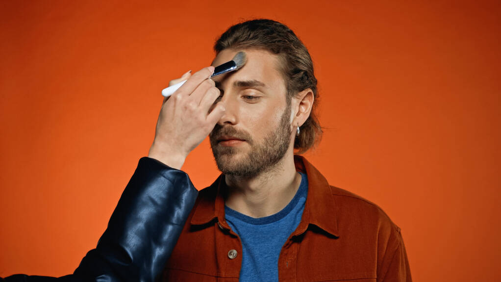 make-up artiest aanbrengen gezicht poeder op jonge man met baard op oranje - Foto, afbeelding