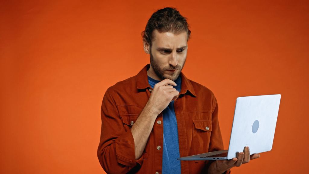 задумчивый молодой фрилансер смотрит на ноутбук на оранжевом  - Фото, изображение