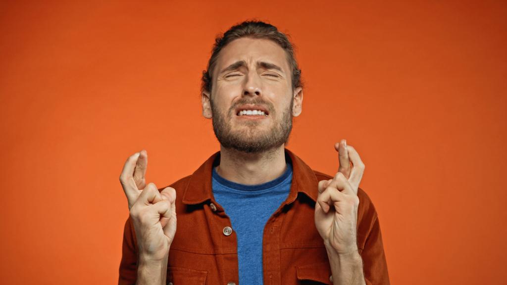 jovem barbudo com olhos fechados e dedos cruzados em laranja  - Foto, Imagem