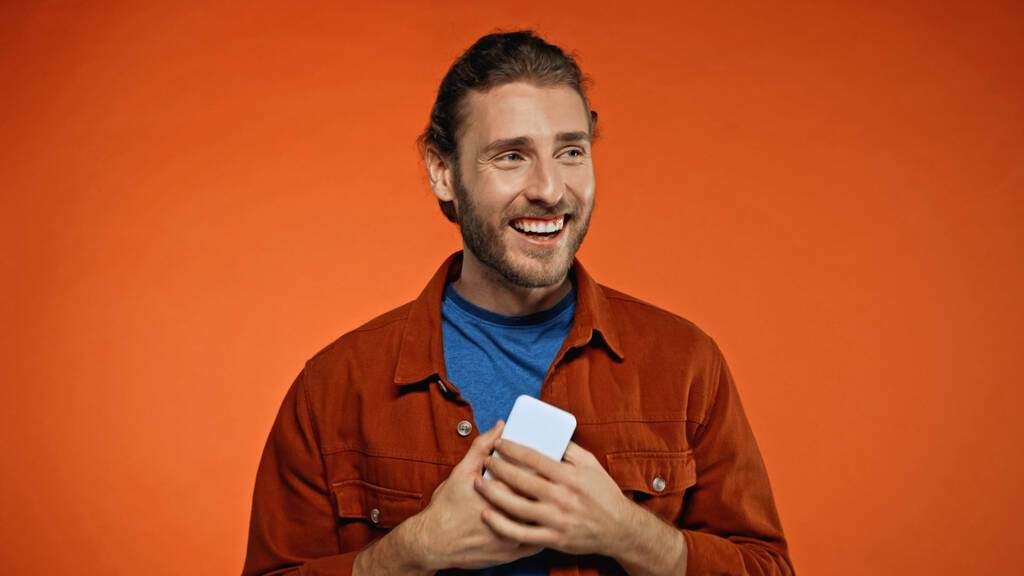 hombre barbudo feliz sosteniendo el teléfono móvil sobre fondo naranja  - Foto, imagen