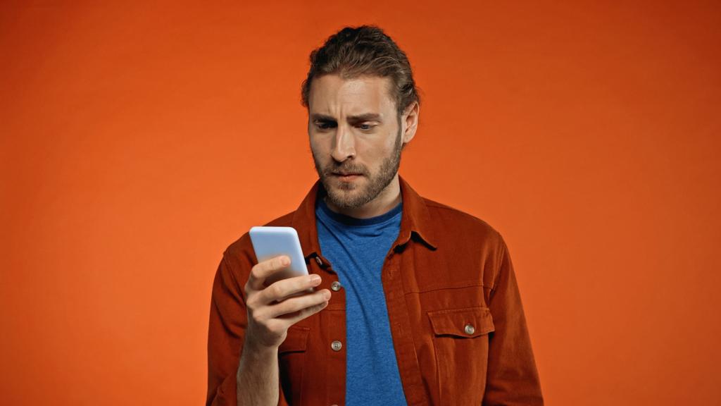 uomo barbuto focalizzato utilizzando il telefono cellulare su sfondo arancione  - Foto, immagini