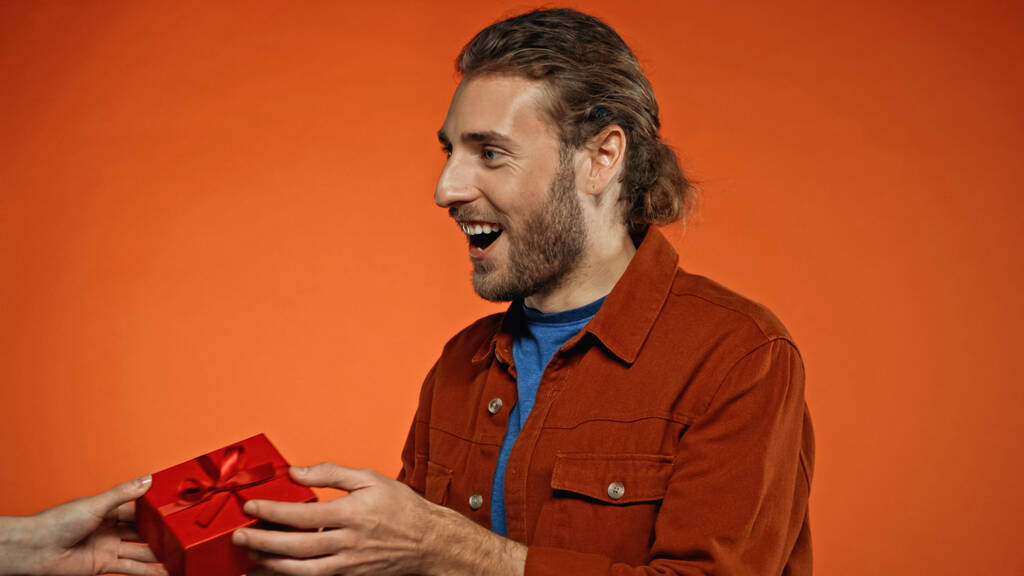 Ευτυχισμένος νεαρός άνδρας που λαμβάνει τυλιγμένο δώρο σε πορτοκαλί  - Φωτογραφία, εικόνα