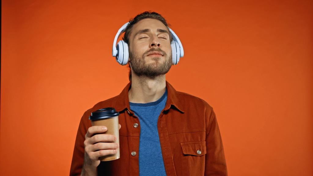 hombre complacido en auriculares inalámbricos sosteniendo la taza de papel y escuchando música en naranja - Foto, Imagen
