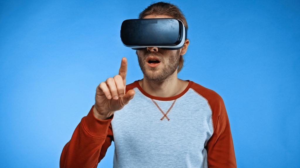bearded young man in virtual reality headset pointing with finger on blue  - Photo, Image