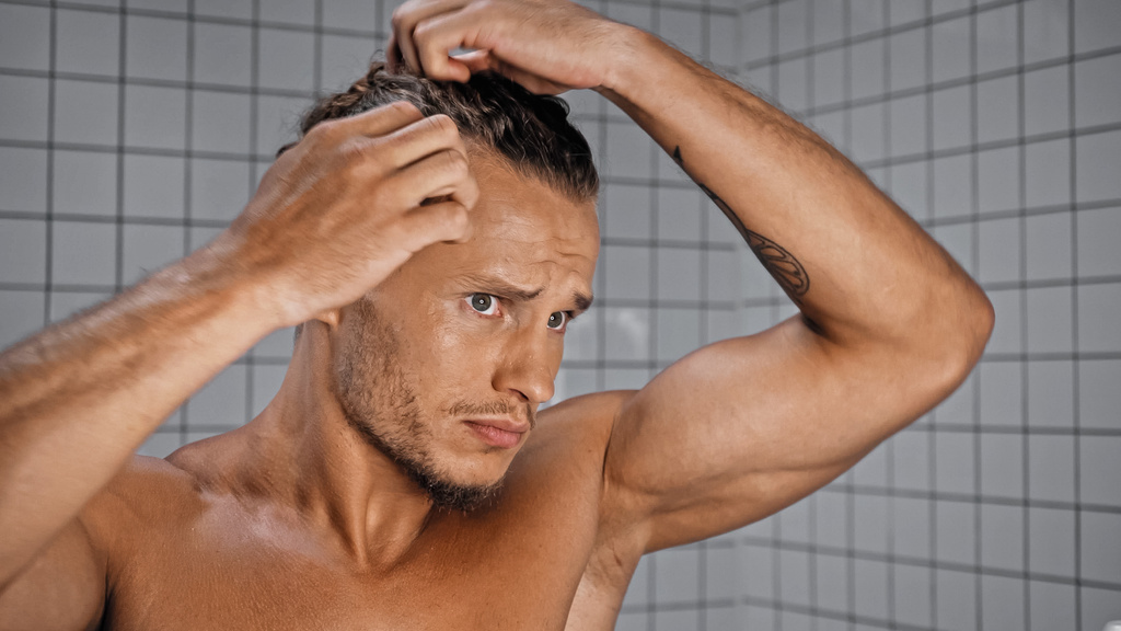 νεαρός άνδρας χωρίς πουκάμισο προσαρμόζει τα μαλλιά στο μπάνιο  - Φωτογραφία, εικόνα