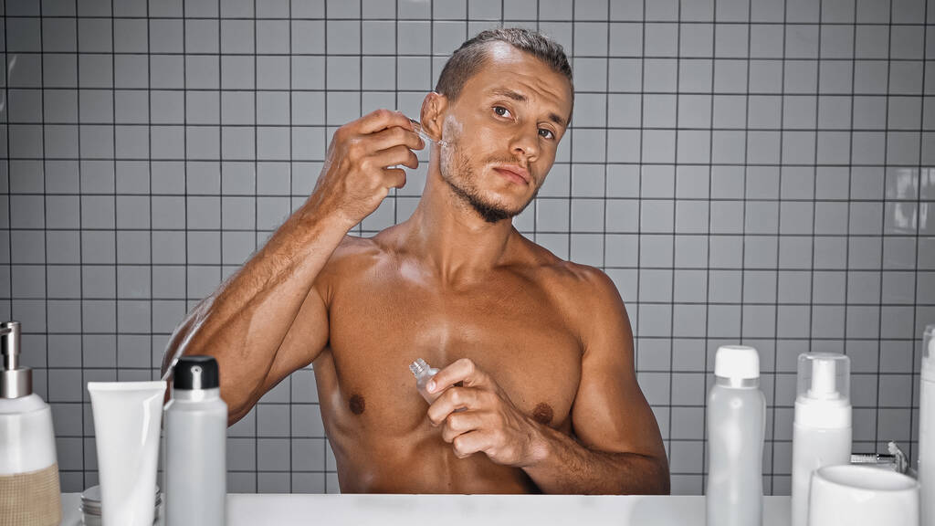 mężczyzna bez koszulki trzymając butelkę i stosując serum w łazience  - Zdjęcie, obraz