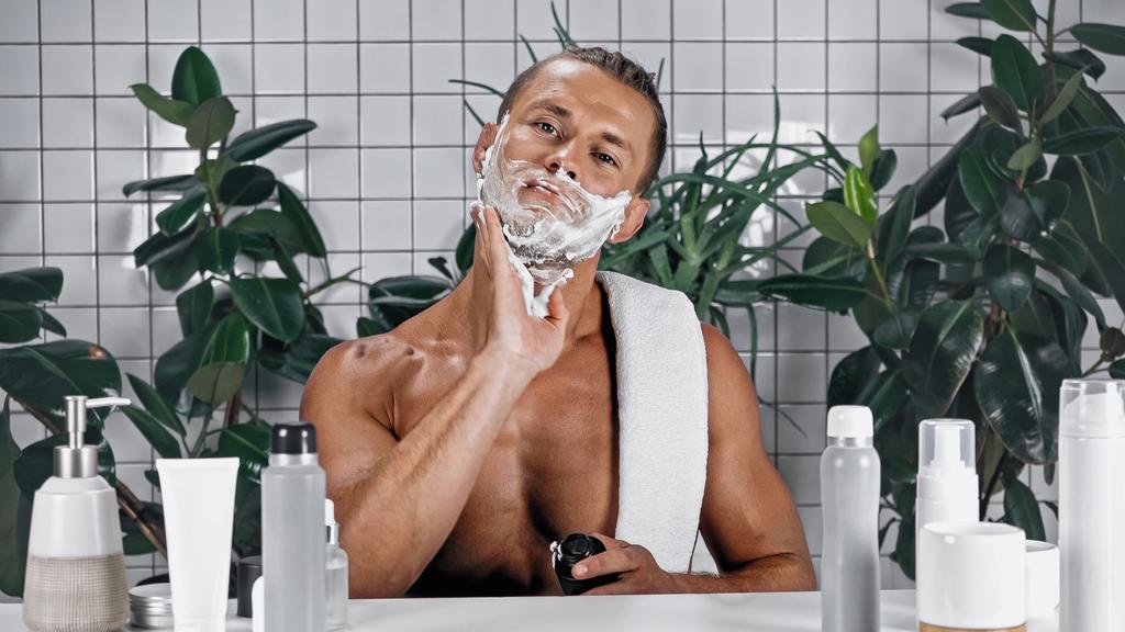 Muž bez košile nanáší pěnu na holení na obličej poblíž lahví a zelených rostlin na rozmazaném pozadí - Fotografie, Obrázek