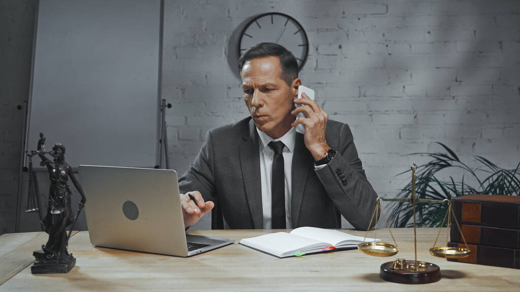 Vakuutusasiamies puhuu älypuhelimella läppärin, kannettavan tietokoneen ja oikeuspatsaan lähellä  - Valokuva, kuva