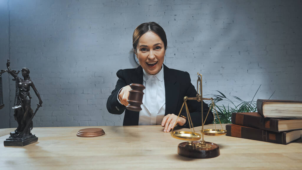Smiling insurance agent holding gavel near books, scales and statuette of justice on table  - Photo, Image