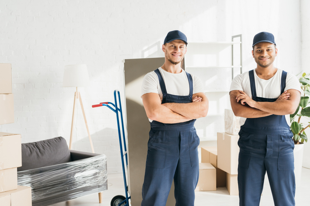 cheerful multicultural movers in caps standing with crossed arms near wrapped furniture  - Photo, Image