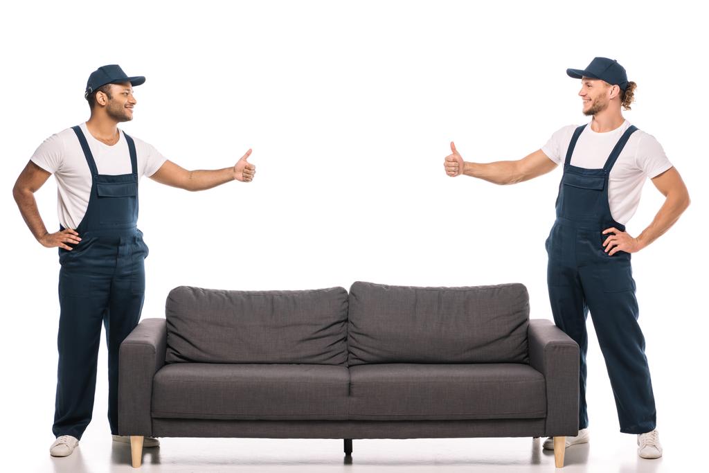 полная длина мультикультурных перевозчиков показывая большие пальцы вверх и глядя друг на друга возле дивана на белом  - Фото, изображение