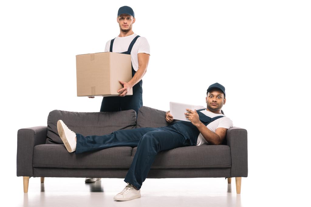 shocked indian man holding digital tablet while resting on sofa near mover with box on white - Photo, Image