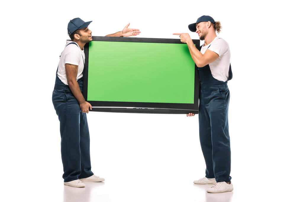 full length of surprised multicultural movers pointing at each other while carrying plasma tv with green screen and on white - Photo, Image