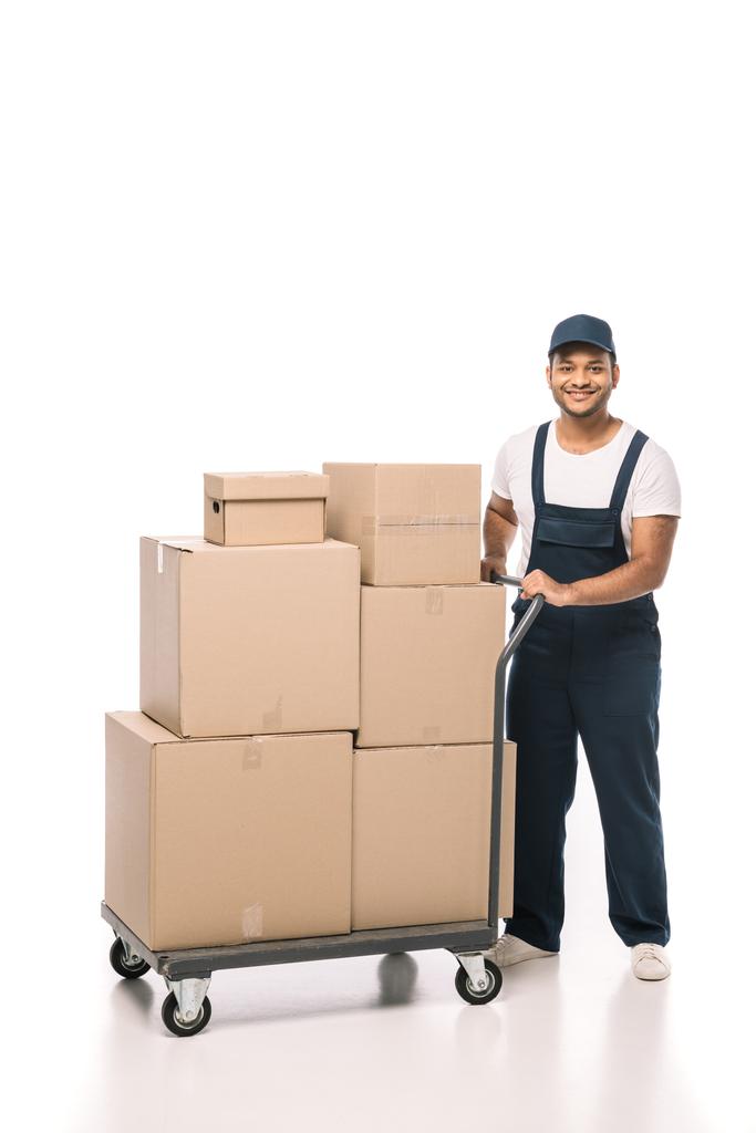 pleine longueur de déménageur indien heureux en salopettes debout près du camion à main avec des boîtes en carton sur blanc - Photo, image