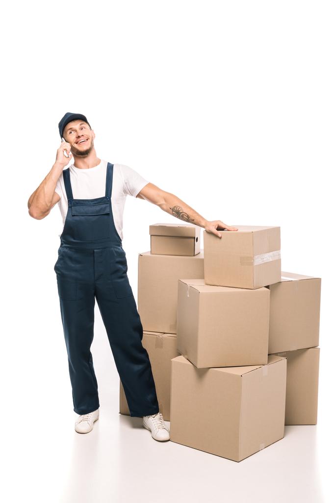 full length of cheerful mover in uniform and cap talking on smartphone near carton boxes on white - Photo, Image