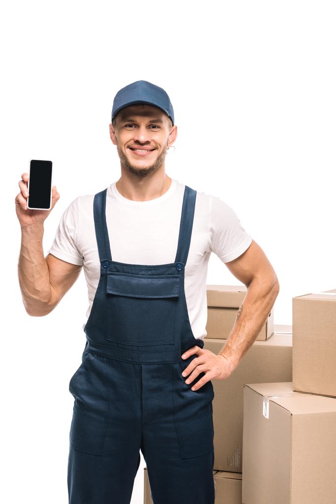 radosny rusznikarz w mundurze trzymający smartfona z czystym ekranem i stojący z ręką na biodrze w pobliżu pudełek kartonowych odizolowanych na biało - Zdjęcie, obraz