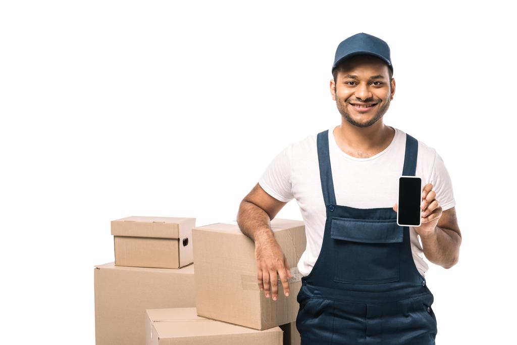 happy indian mover in overalls holding smartphone with blank screen near carton boxes isolated on white - Photo, Image