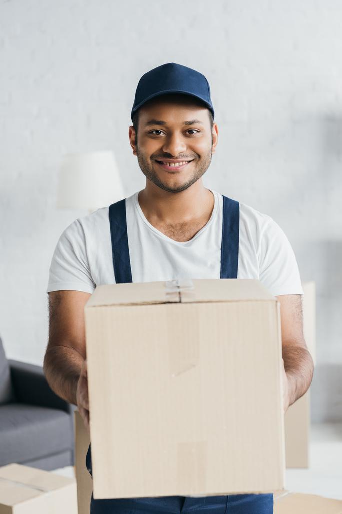 positive indian courier in cap and uniform holding box in apartment  - Photo, Image