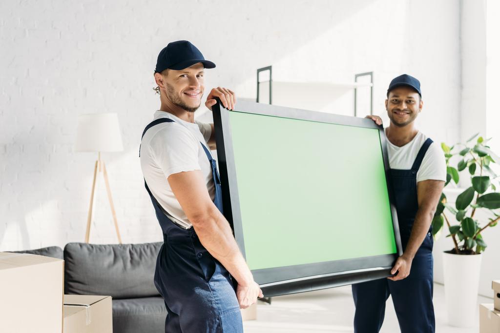 feliz multicultural movers en uniforme llevando plasma tv con pantalla verde en apartamento  - Foto, Imagen