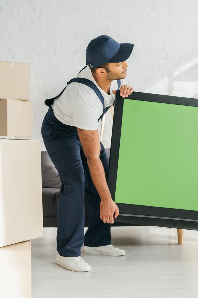 Indischer Mover in Uniform trägt Plasma-TV mit grünem Bildschirm in der Wohnung  - Foto, Bild