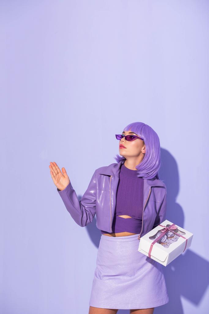 jonge vrouw gekleed in pop stijl met cupcake doos op violette kleurrijke achtergrond - Foto, afbeelding