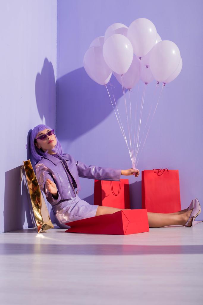 młoda kobieta ubrana w stylu lalki siedzi z torbami na zakupy i balonami na fioletowym tle - Zdjęcie, obraz